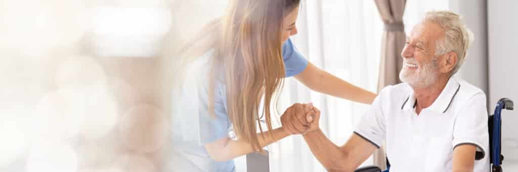 Eine freundliche Pflegekraft lächelt und hält die Hand eines älteren Mannes im Rollstuhl. Die Atmosphäre wirkt warm und einfühlsam und vermittelt das Gefühl von Fürsorge und Unterstützung
